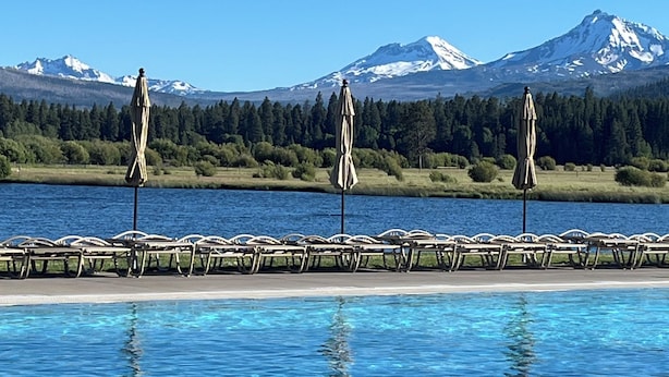 Una piscina con sillas de descanso y sombrillas, un cielo azul despejado y una cordillera de montañas al fondo.