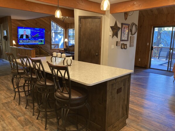 A kitchen with a large island, bar stools, a television displaying financial news, and a wooden interior. - Black Butte Home- Best Deal on the Ranch! (Black Butte)