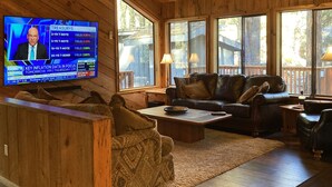 Una sala de estar acogedora con paredes y techo de madera, un televisor mostrando noticias financieras, y una zona de estar confortable con un sofá y sillones.