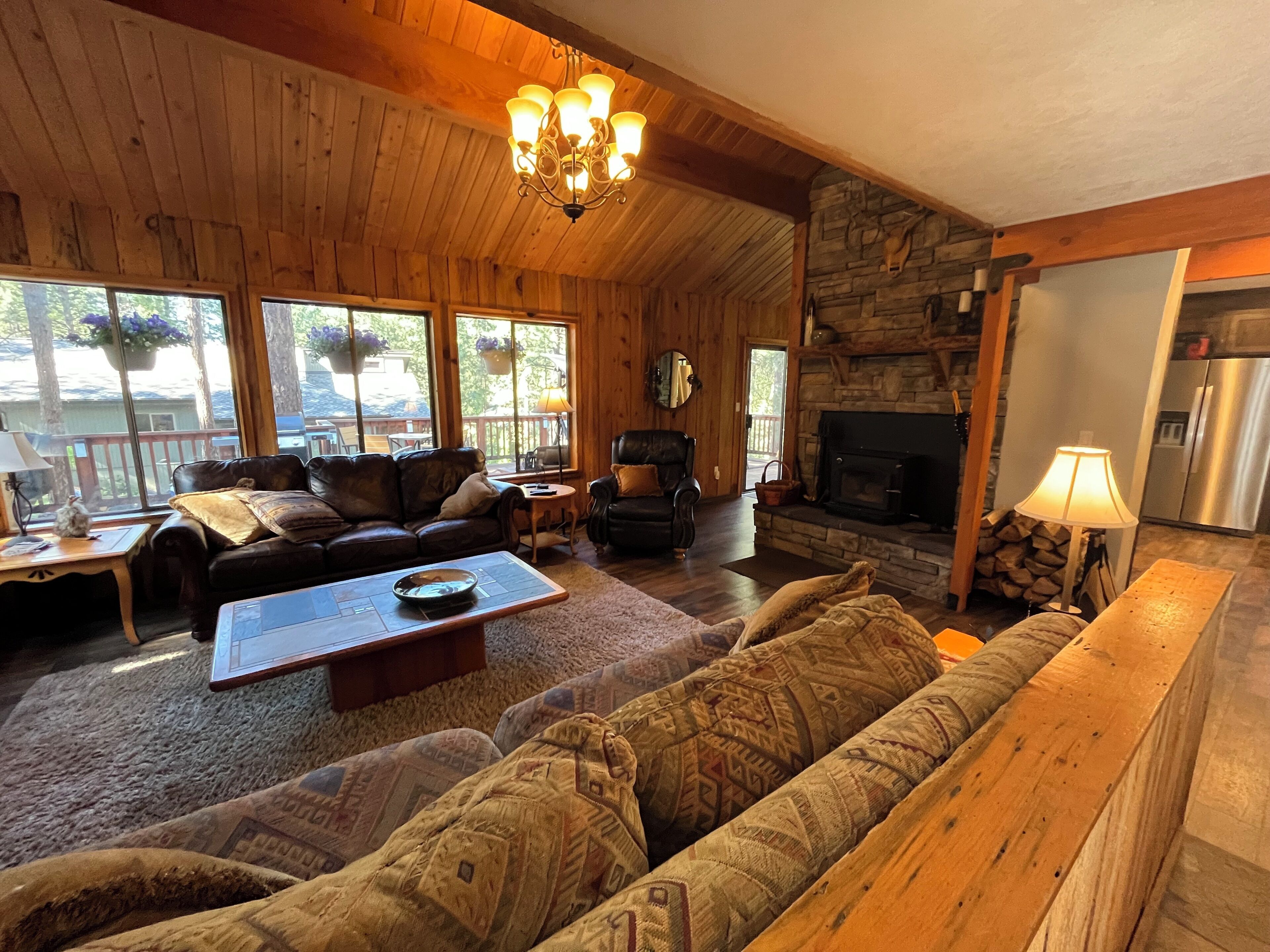 A cozy cabin interior with a stone fireplace, wooden walls, and a large window overlooking a deck.