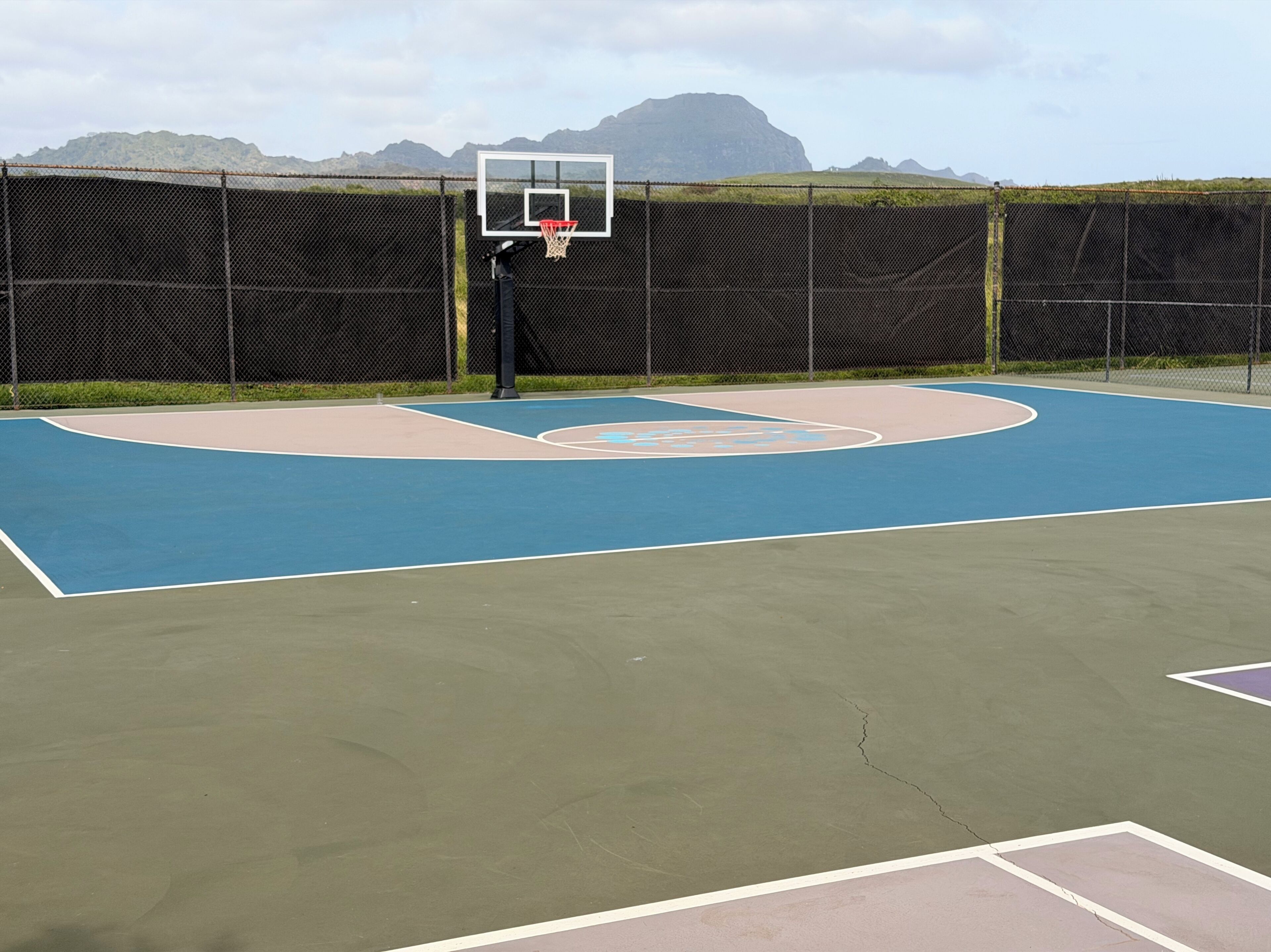 Ein Basketballplatz im Freien mit einem Maschendrahtzaun, einem Basketballkorb und einer Bergkulisse.