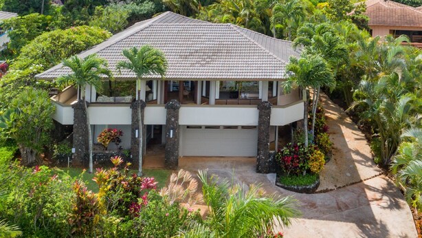 Une maison à deux étages, dotée d’un toit en tuiles, entourée d’une végétation tropicale luxuriante et d’un jardin bien entretenu.