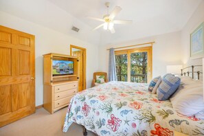 Ein Schlafzimmer mit einem Bett, einer Holzkommode, einem Fernseher darauf, einem Deckenventilator und einem Fenster mit Blick auf Bäume.
