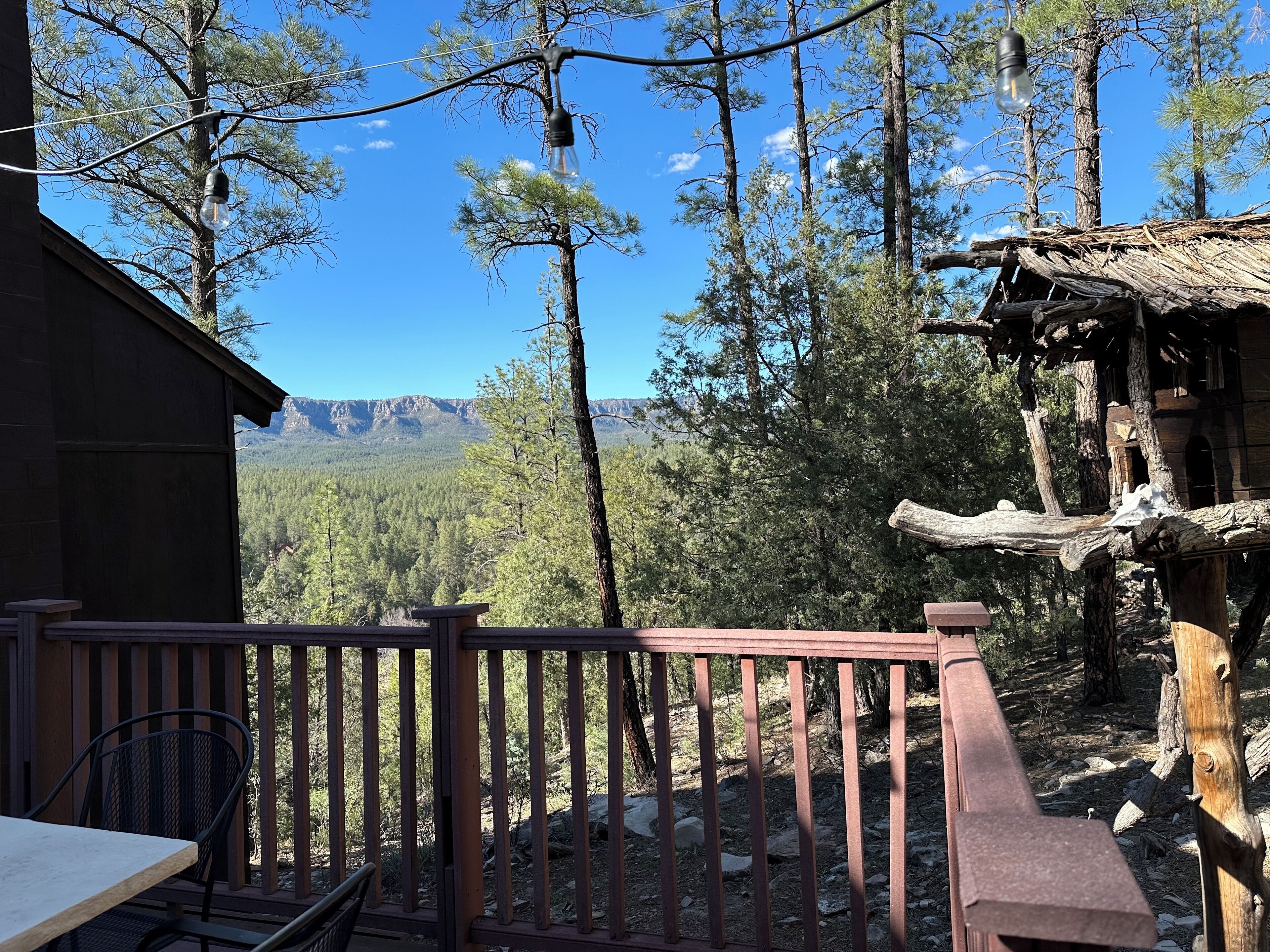 A wooden deck with a table and chairs, overlooking a forested area with mountains in the distance.