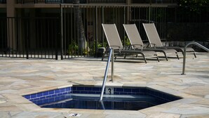 Una piscina con una escalera metálica, dos sillas de descanso y un suelo de baldosas de piedra.