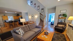 Ein geräumiges Wohnzimmer mit einem grauen Sofa, einem Holztisch und einem gemusterten Teppich. Eine Treppe führt zu einem Balkon mit Blick.