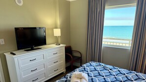 A hotel room with a bed, a TV, a dresser, a chair, and a window with a view of the sea.