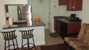 A compact kitchen with a refrigerator, a coffee maker, and a sink. There are two bar stools and a patterned sofa in the living area. - Mardi Gras Cottage, 4 km to French Quarter (ARABI)