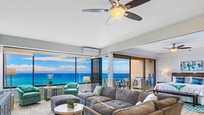 Een moderne woonkamer met een grote bank, een salontafel en uitzicht op de oceaan door grote ramen. Achtergrond is een bed met een blauw-witte dekbedovertrek.