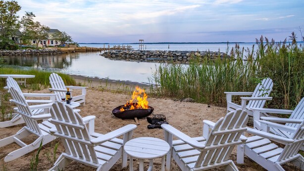 A beachside setting with a fire pit and white Adirondack chairs, overlooking a calm body of water and a distant shoreline. - 240' Private Beachfront @Kent Island Love Point - Great for Families (stevensville)