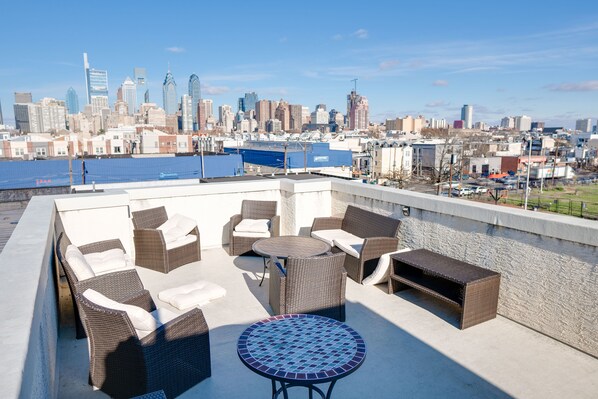 A rooftop patio with wicker furniture and a mosaic table, overlooking a city skyline and waterfront.