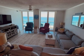 A living room with a large TV, a sofa, two armchairs, a coffee table, and a view of the ocean through large windows. - Great Family Retreat w Amazing Views. #11D (Pensacola Beach)