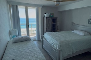 A bedroom with two beds, a window with a view of the beach, a ceiling fan, and a small cabinet. - Great Family Retreat w Amazing Views. #11D (Pensacola Beach)