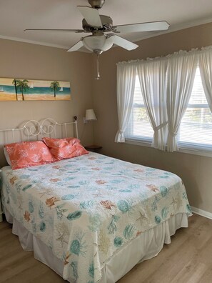 A bedroom with a bed, a ceiling fan, a window with curtains, a nightstand with a lamp, and a painting on the wall.