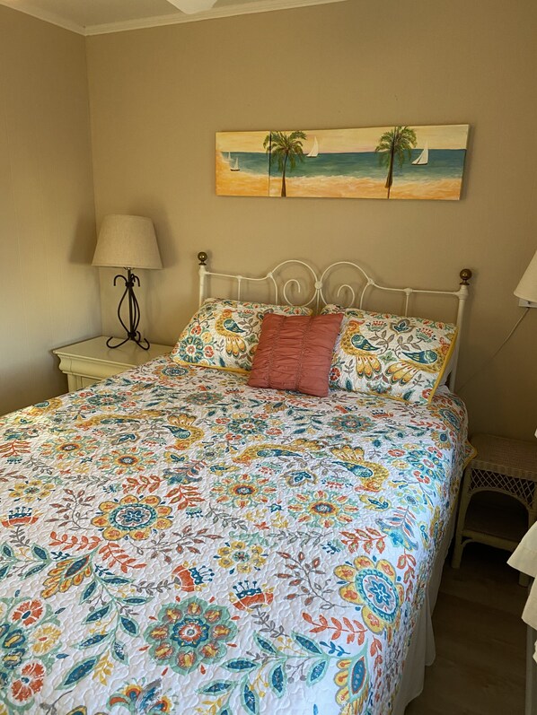 A bedroom with a bed, a nightstand, a lamp, a painting of a beach scene, and a pet carrier.