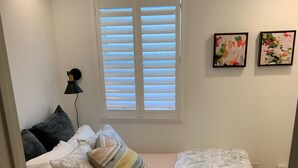 Un dormitorio con cama, almohadas, mesita de noche, lámpara, ventana con persianas y dos cuadros en la pared.