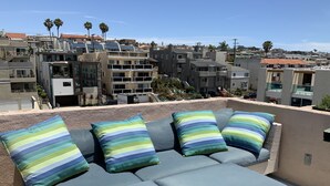 Un área de lounge en la azotea con un conjunto de sofás y cojines, con vistas a un vecindario residencial con edificios modernos y palmeras.