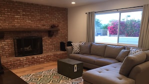 A living room with a brick fireplace, a sectional sofa, a coffee table, and a large window with a view of a garden.