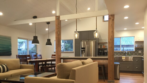 A modern living room with a sofa, coffee table, and pendant lights. A kitchen with a stainless steel refrigerator and a dining area with a table and chairs are visible.