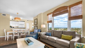 A living room with a sofa, a dining table with chairs, a kitchen area, and large windows with blinds. - Beach Colony West 4A- Gulf-front 3 BD/3BA w/Beach Service & Sleeps up to 8 (Navarre Beach)