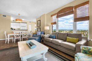 Una sala de estar con un sofá, una mesa de comedor con sillas, una zona de cocina y ventanas amplias con persianas.