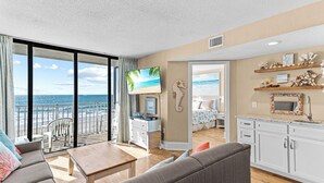 A spacious living room with a large TV, a balcony with a view of the ocean, and a kitchen area with white cabinets and decorative items. - Oceanfront Condo, Gorgeous, Clean & Remodeled located at Sands Beach Club #325 (Myrtle Beach)