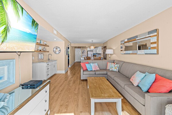 A modern living room with a flat-screen TV, a grey sofa, wooden flooring, and a kitchen in the background.