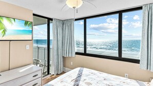 A bedroom with a large window, a ceiling fan, a bed with a beach-themed bedspread, a dresser, and a chair by the window. - Oceanfront Condo, Gorgeous, Clean & Remodeled located at Sands Beach Club #325 (Myrtle Beach)