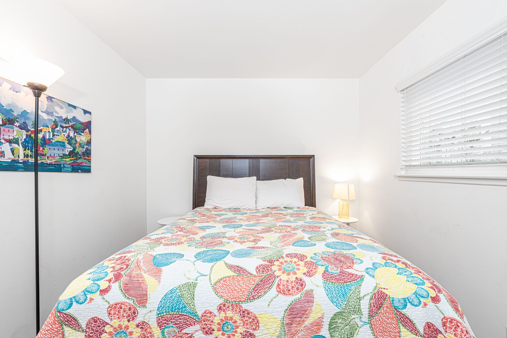 A bedroom with a bed, a nightstand, a lamp, a painting, and a window with blinds.