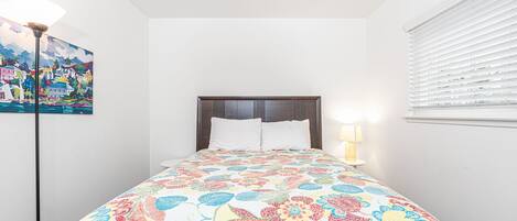 A bedroom with a bed, a nightstand, a lamp, a painting, and a window with blinds.