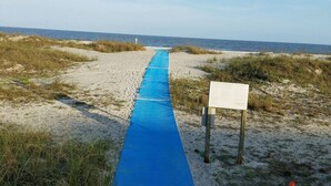 Un sendero arenoso en la playa con un tapete azul que lleva al mar, una pasarela de madera y un cono de tráfico.