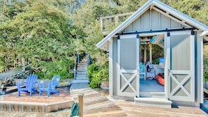A wooden deck with blue Adirondack chairs, a covered outdoor area with a bench, and a staircase leading to a building with a gabled roof. - Eastern Lake Lakefront! Easy access to the Gulf! Pirate Ship & Treehouse! (Santa Rosa Beach)