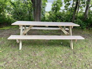 Una mesa de picnic de madera con banco, ubicada en un área verde con árboles y una casa al fondo.