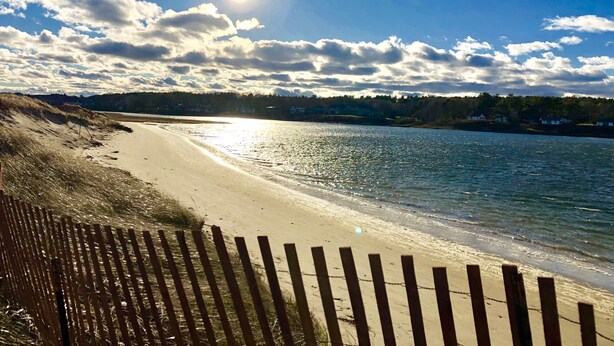 Ein Strand mit einer Holzzahlung im Vordergrund, einem sandigen Ufer und ruhigem Wasser.