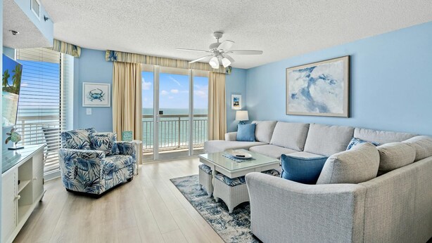 A living room with a sofa, armchairs, a coffee table, and a ceiling fan. There is a view of the ocean through large glass doors. - Beach Chic Oceanfront Condo, Jacuzzi Tub, Lazy River | Crescent Shores - N 1203 (North Myrtle Beach)