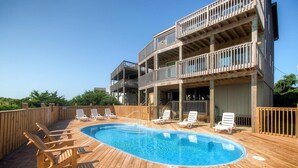 A wooden deck with a pool, lounge chairs, and a multi-story building with balconies. - Oceanfront Home in Avon with Private Saltwater Pool, Hot Tub, and Tiki Bar (Avon)