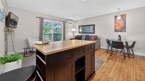 Een moderne keuken met een centraal eiland, een eetgedeelte met een glazen tafel en stoelen, en uitzicht op de omgeving door grote ramen.