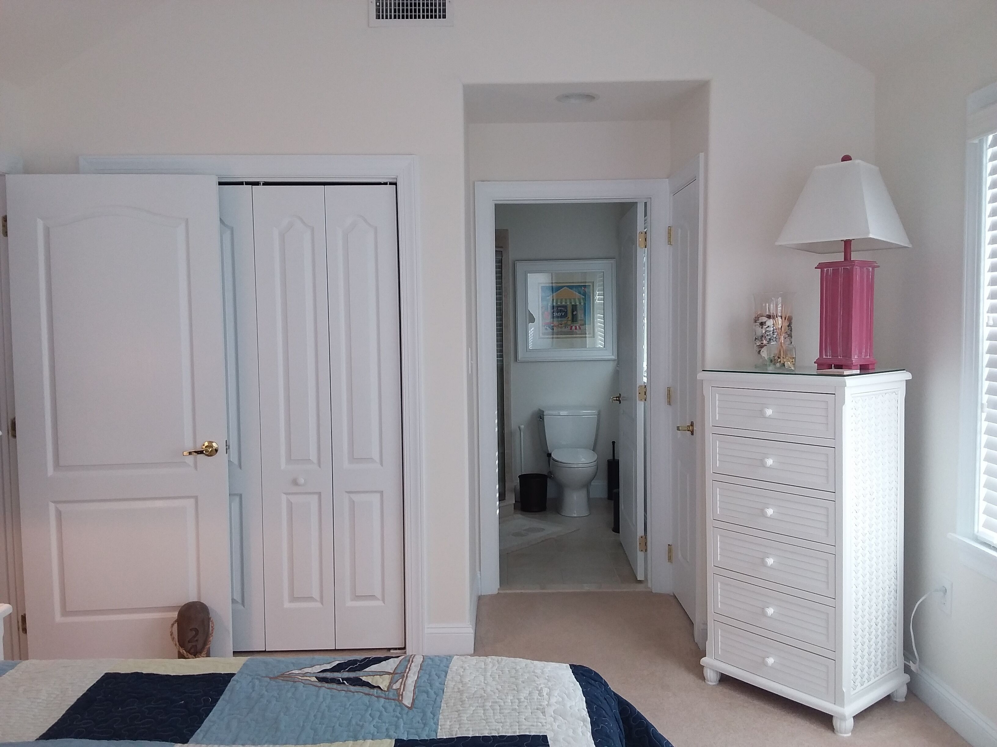 A bedroom with a bed, a dresser, a lamp, and a door leading to a bathroom.