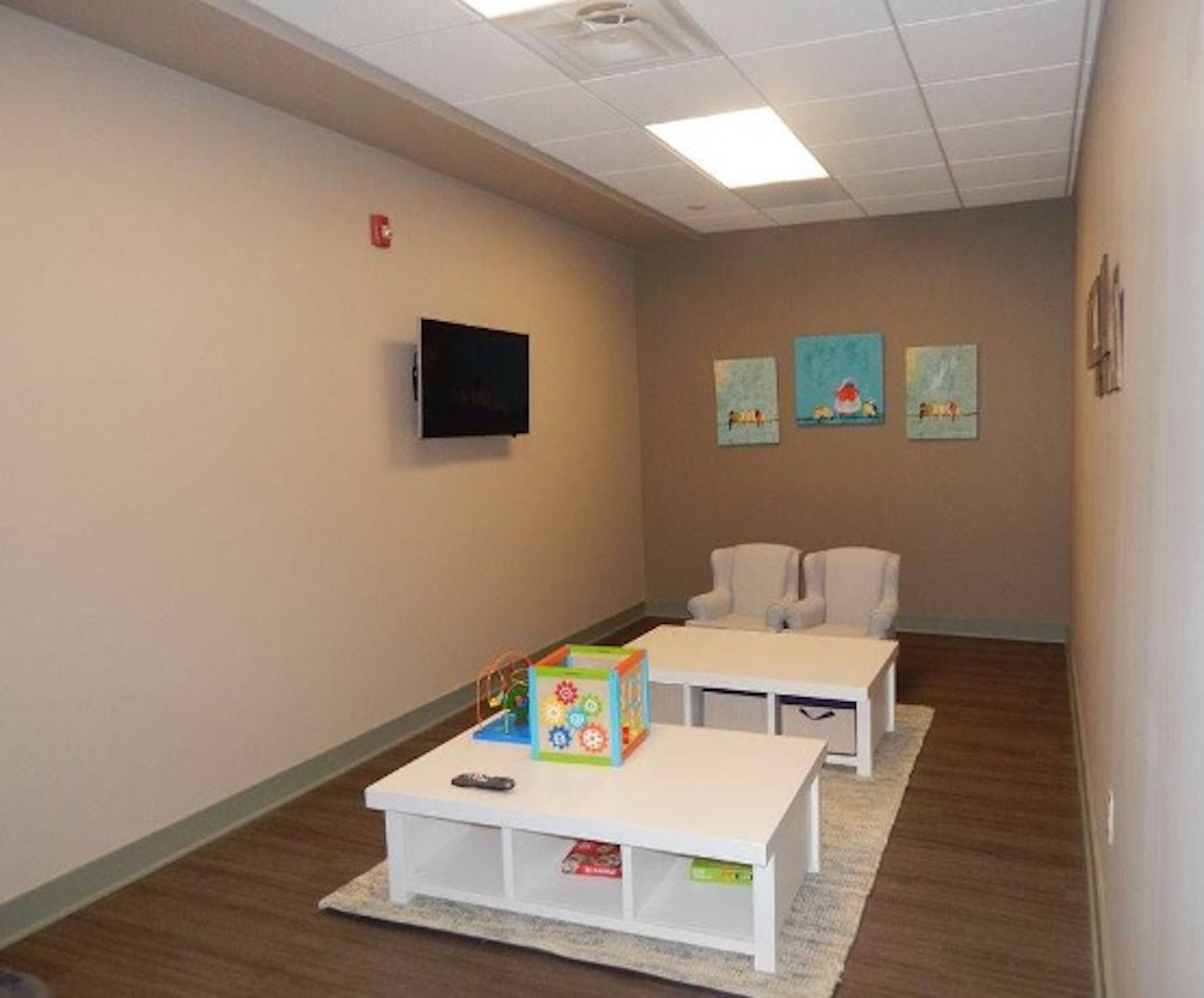 Una habitación con dos mesas blancas, un televisor en la pared y tres cuadros enmarcados.