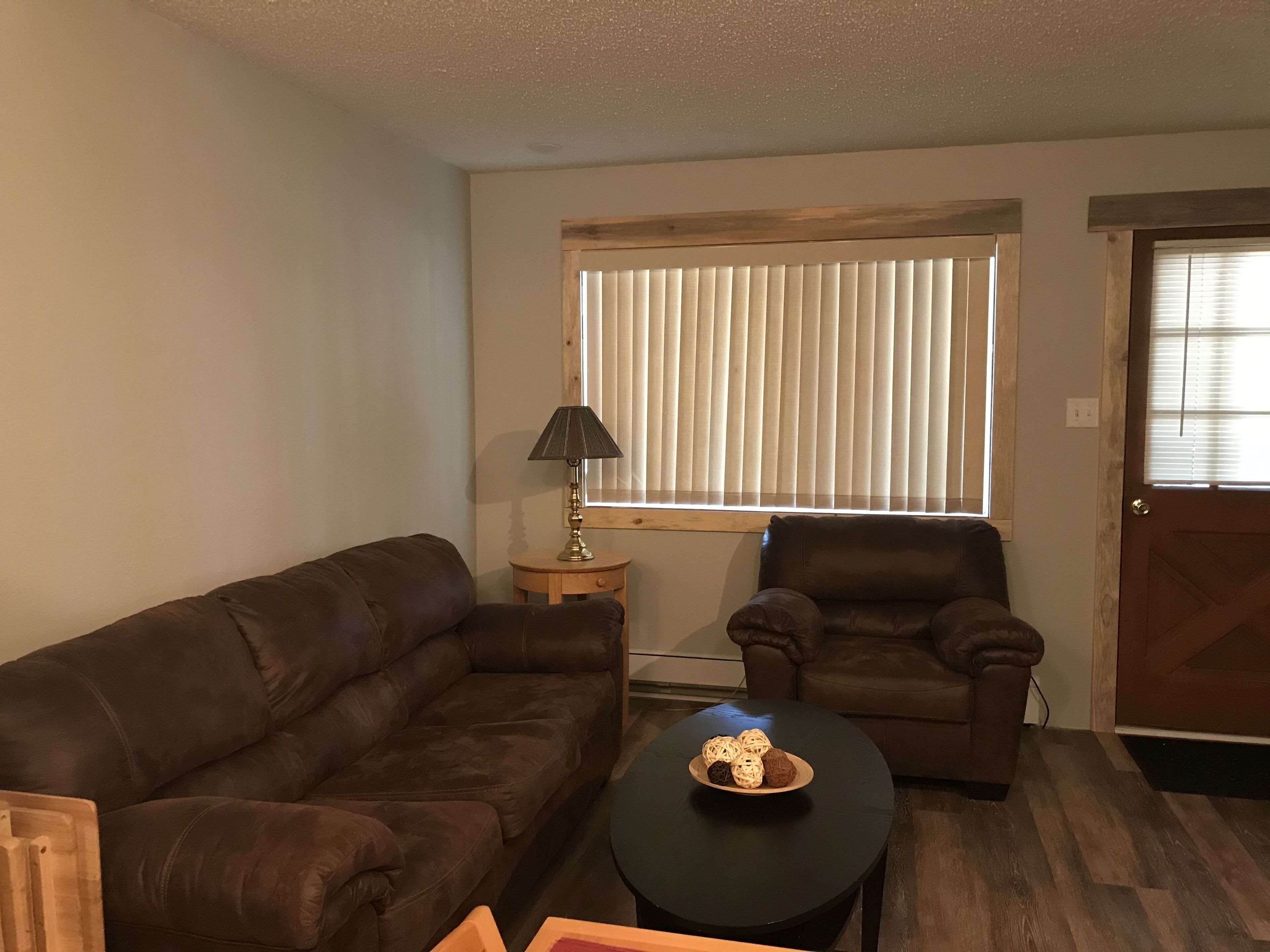 A living room with a brown sofa, a brown armchair, a small round table with a lamp, and a window with blinds.