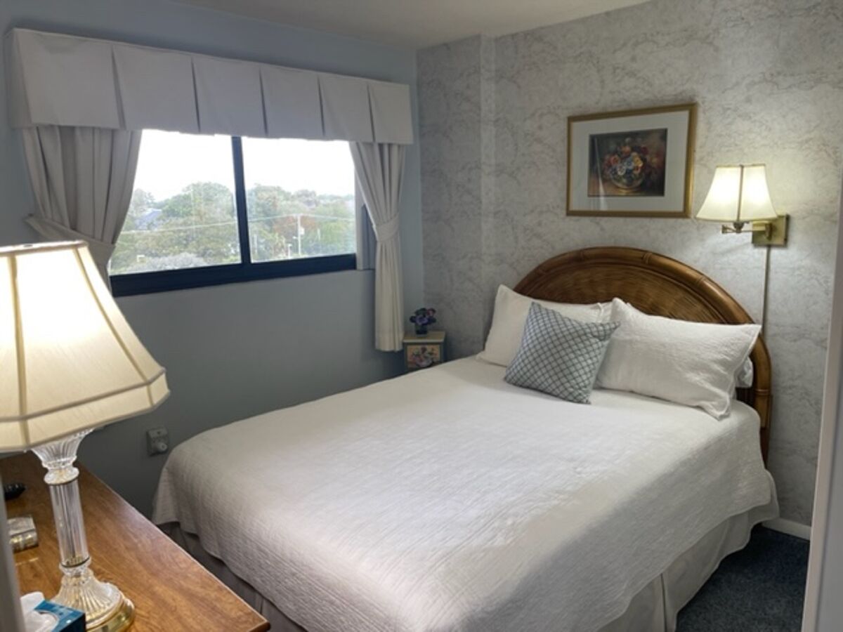 A neatly made bed with white linens, a wooden headboard, and a bedside lamp. A window with curtains is visible, and a framed picture hangs on the wall.