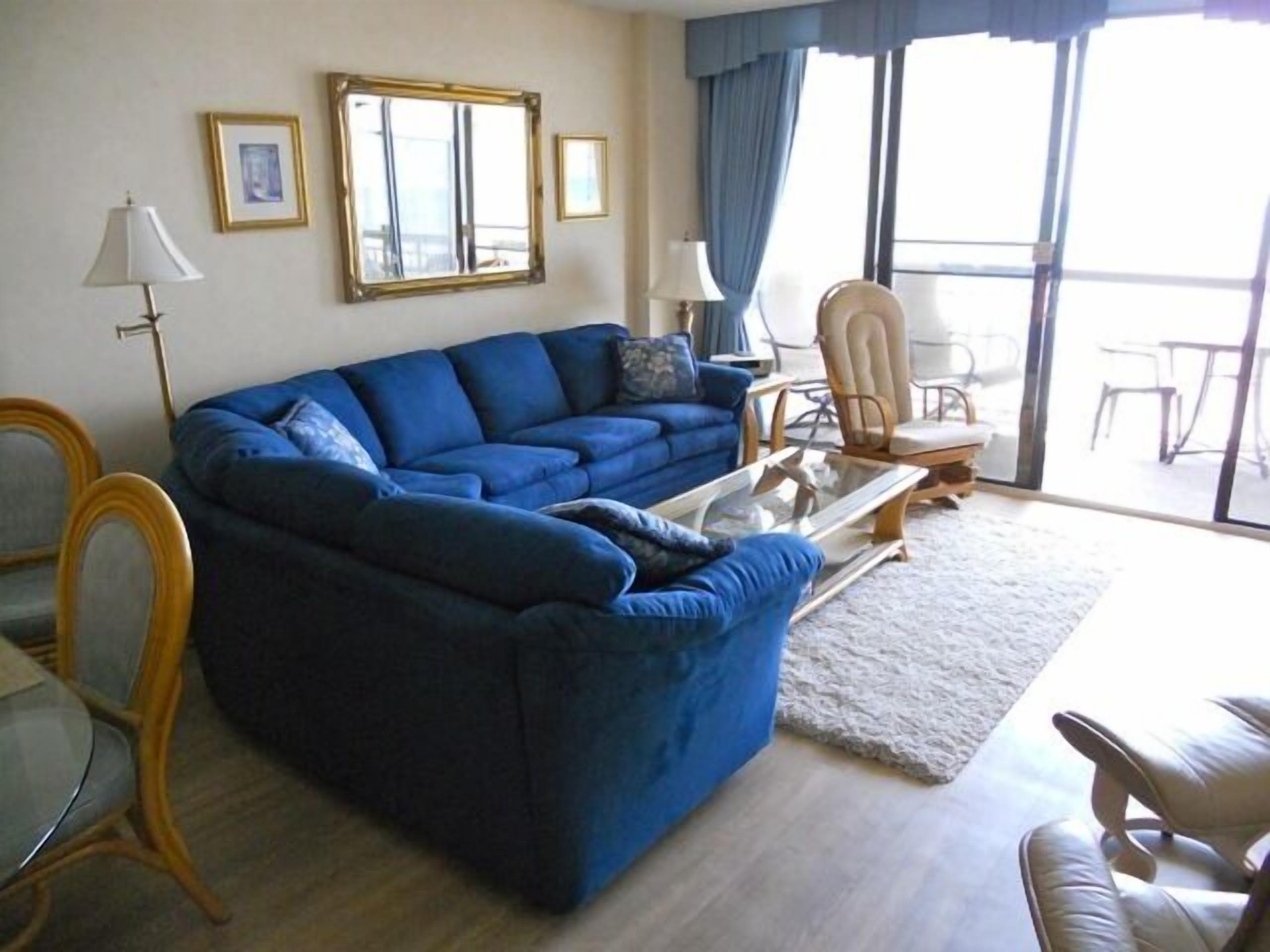 Un salon avec un canapé bleu, une table basse en verre, un fauteuil blanc et une table à manger avec des chaises.