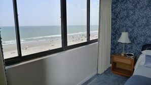 Une chambre avec une grande fenêtre donnant sur une plage, une table de chevet avec une lampe et un lit recouvert d’une couverture bleue.