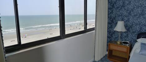 A room with a large window overlooking a beach, a bedside table with a lamp, and a bed with a blue cover.
