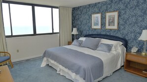 Une chambre avec un grand lit, deux tables de chevet avec des lampes, un fauteuil et une vue sur la mer par une grande fenêtre.