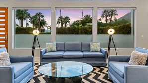 Un salón moderno con un sofá azul, dos sillones, una mesa de centro de vidrio y grandes ventanas con vistas a palmeras y una montaña.