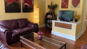 A living room with a leather couch, a glass coffee table, a bookshelf, a TV on a stand, and artwork on the walls.