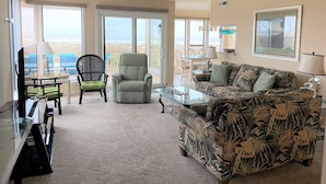Amplia sala de estar con un gran ventanal con vista a la playa, un sofá confortable, una mesa de comedor y un televisor.