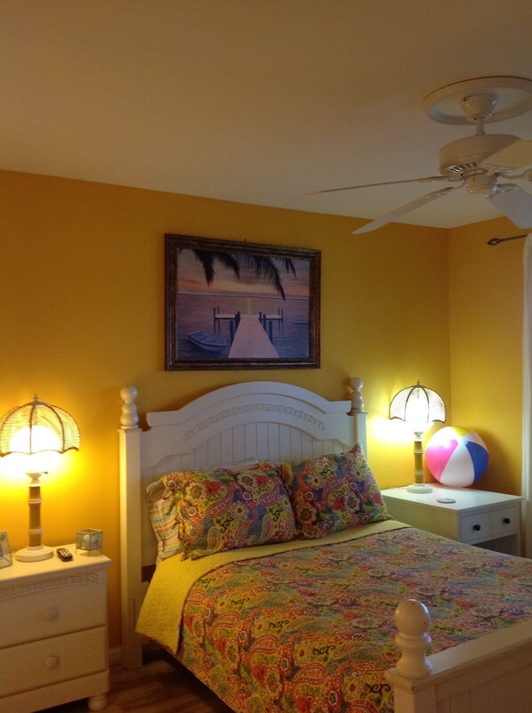 A bedroom with a bed, nightstands, a lamp, a beach painting, and a ceiling fan.