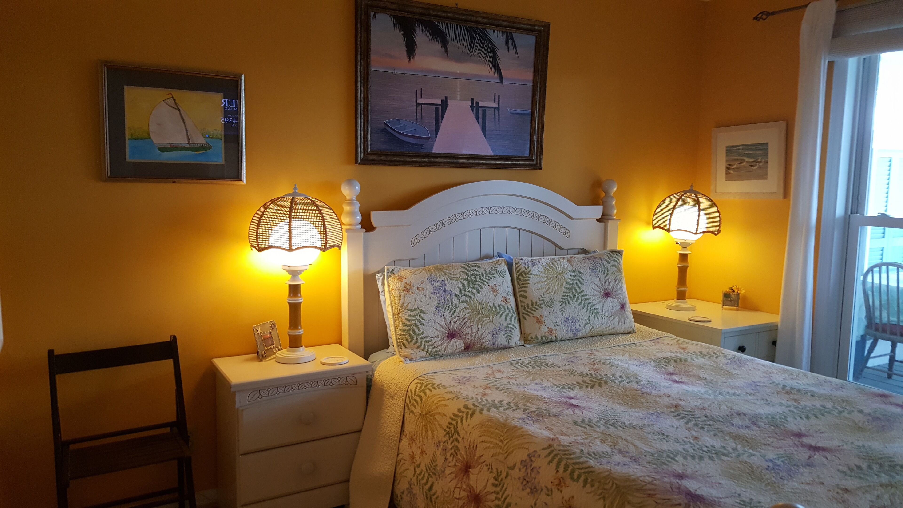 A bedroom with a bed, two bedside tables, two lamps, a chair, and two framed artworks on the wall.